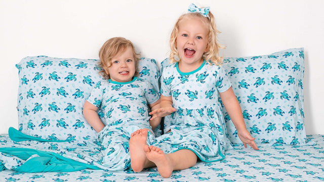 Two laughing kids sitting up in a big-kid bed made with sea turtle print sheets and pillowcases