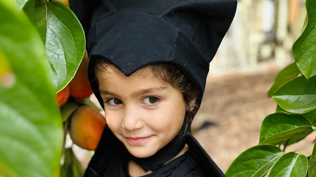Toddler in a black bat costume with hood and ears outdoors among citrus leaves
