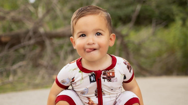 Toddler crouching outdoors in short pajamas, sticking out his tongue with a cheeky grin