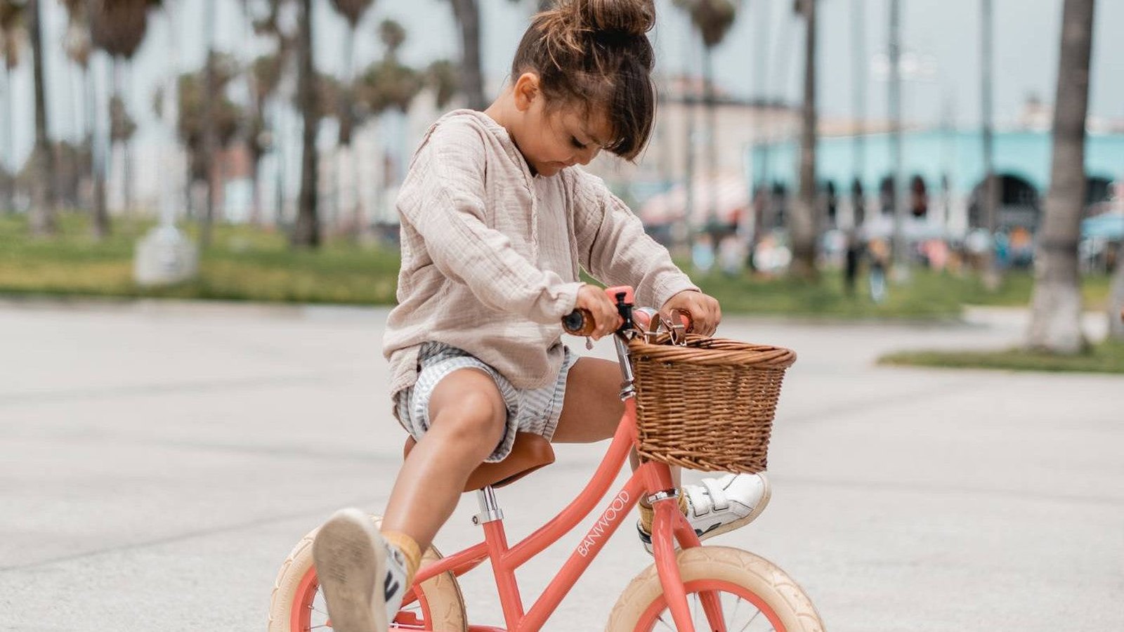 Toddler riding a coral Banwood balance bike with a wicker front basket along a palm-lined seafront path