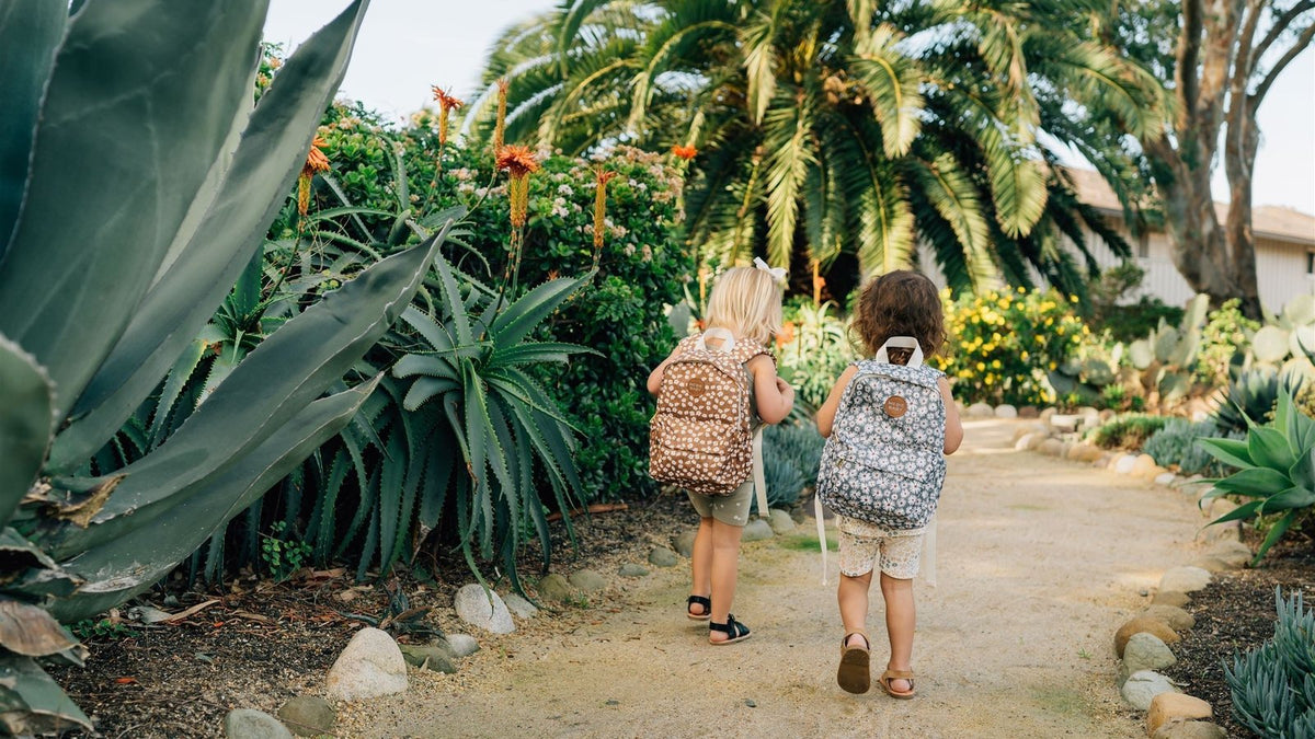 Two toddlers wearing mini backpacks walking hand in hand down a palm-lined garden path