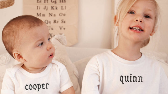 Baby and older sister sitting side by side in white tees embroidered with their names, Cooper and Quinn, below an alphabet wall chart