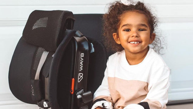 Smiling toddler sitting on a driveway beside a folded WAYB Pico travel car seat