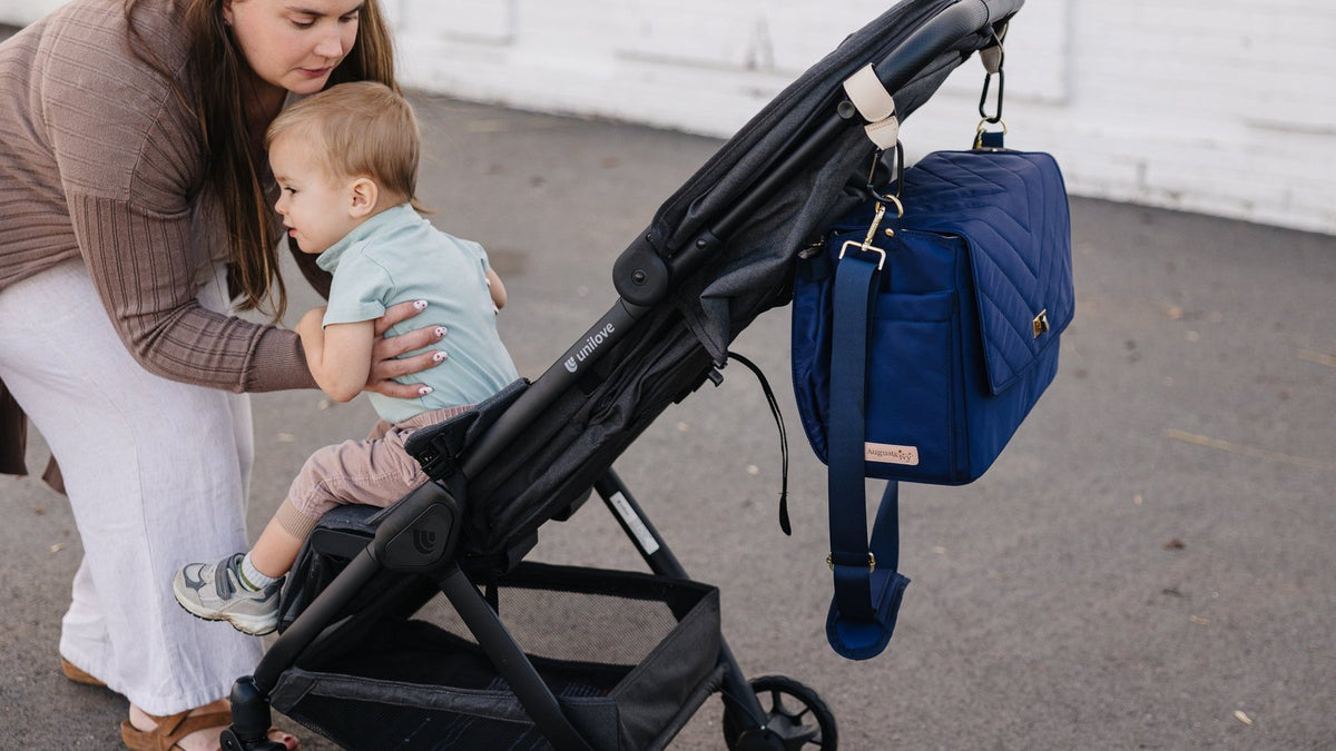 Mother lifting her toddler into a stroller with a navy diaper bag clipped to the handlebar