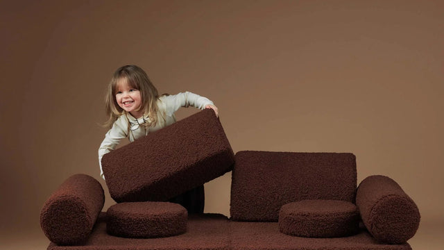 Little girl stacking the cushions of a Wigiwama Warm Hazel teddy-boucle Settee play couch into a fort