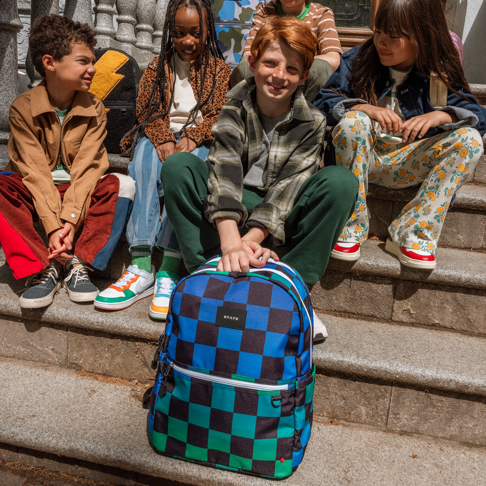 Group of school-age children sitting on steps together with a blue and green checkered backpack