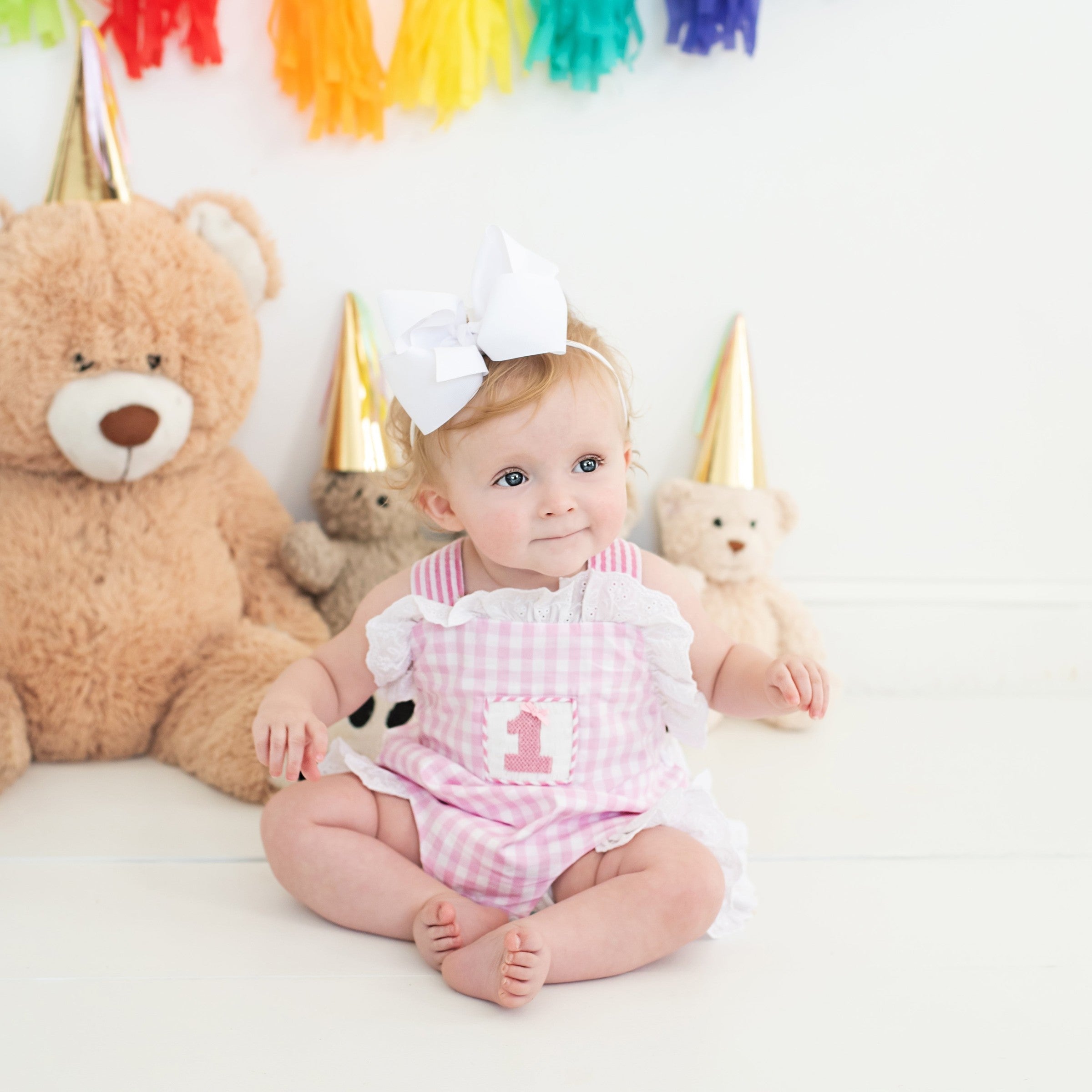 Baby girl in a pink gingham first birthday romper with party hats and teddy bears